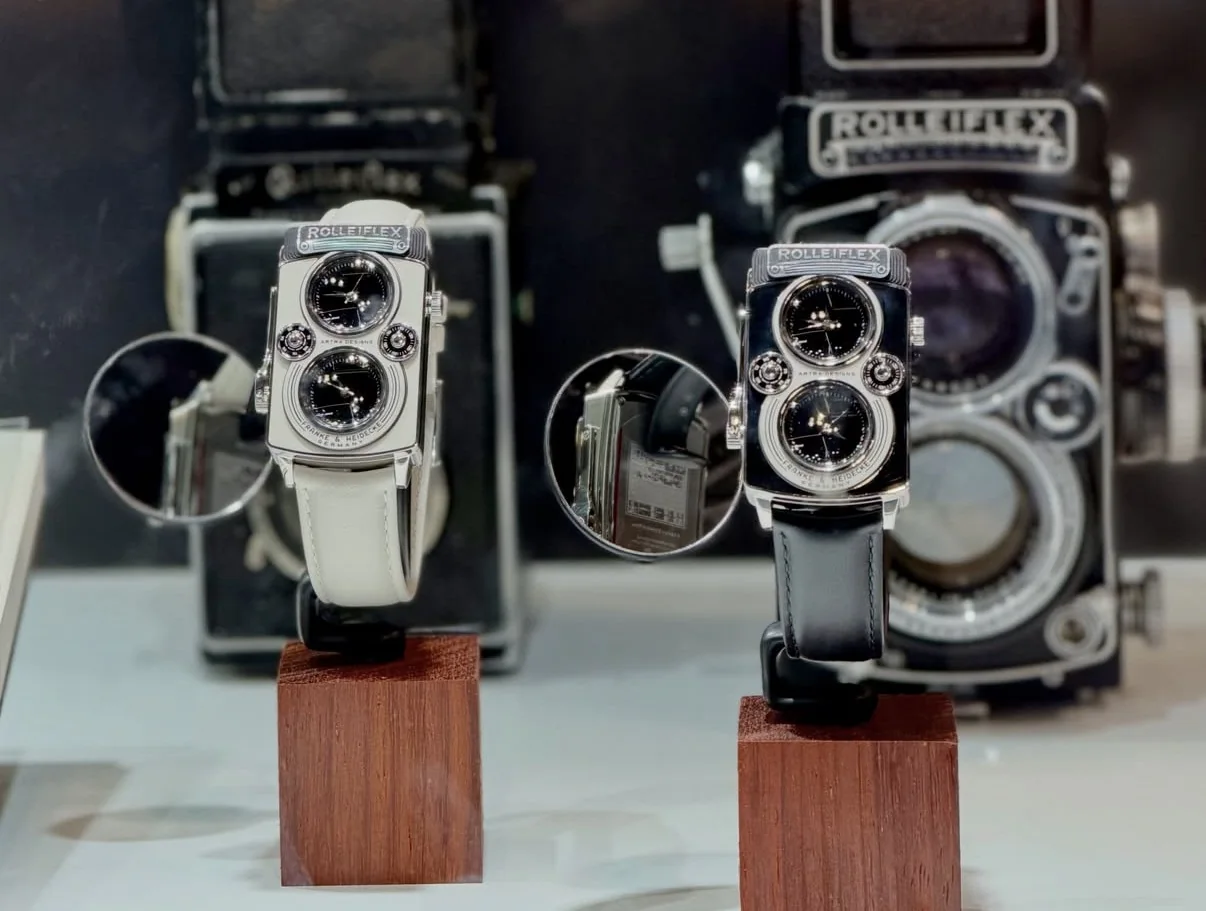 전시대에 놓인 CAMERA DUAL ROLLEIFLEX 두 점. 왼쪽은 흰 가죽 스트랩, 오른쪽은 검은 가죽 스트랩이고 뒤로 실제 롤라이플렉스 카메라 두 대가 서 있다