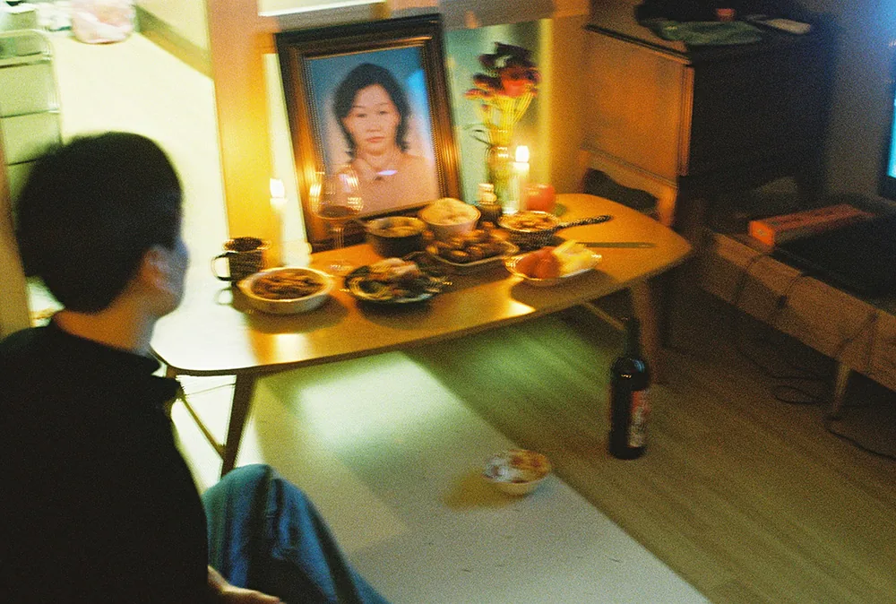 방 안에 차린 제사상. 낮은 좌탁 위에 영정 사진과 촛불, 꽃병, 나물과 과일이 놓여 있고, 왼쪽 앞에 검은 옷을 입은 사람이 흐릿하게 앉아 있다. 바닥에는 소주병과 그릇이 하나씩 있다.