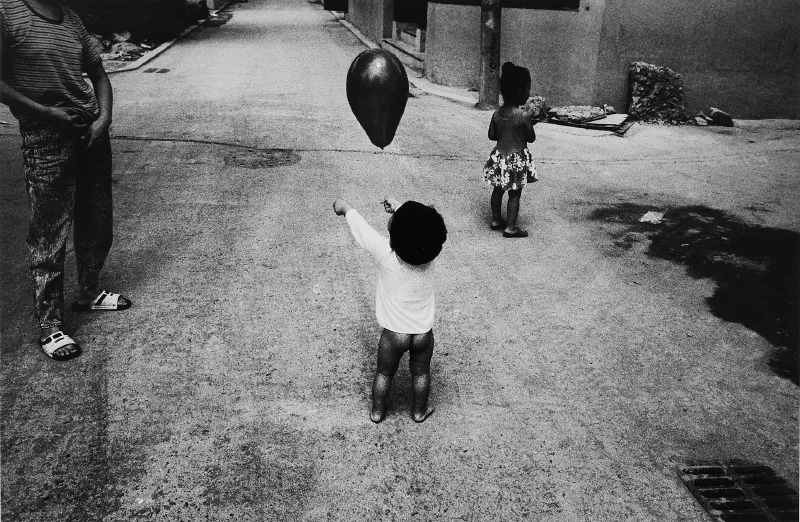 狭い路地のモノクロ写真。ほとんど裸足の幼い子どもが背を向けて黒い風船に向かって腕を伸ばし、手前にもうひとりの子どもが立っている。左端にはサンダルを履いた大人の脚が切れて入り込んでいる。
