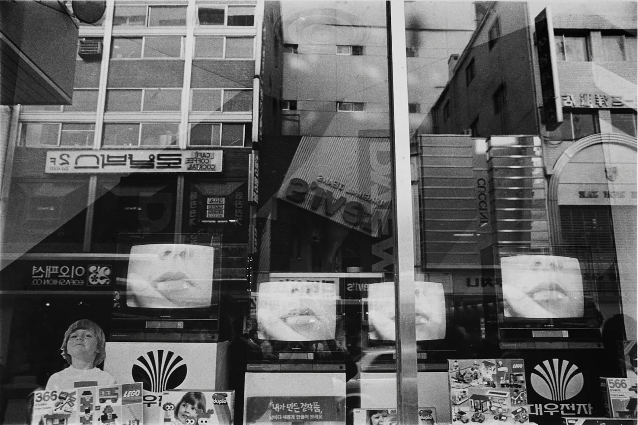 店のショーウィンドーに映る都市を撮ったモノクロ写真。陳列された何台ものテレビに同じ唇のクローズアップが映り、ガラスには向かいの建物と看板が重なって映り込んでいる。左下では子どもがひとり、ショーケースの前に立っている。