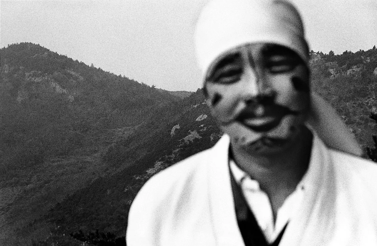 モノクロ写真。白い頭巾に白い服の人物が、顔に黒い塗料で眉とひげ、頬のほくろを描き、カメラのすぐ近くに立っている。顔はピントが外れてぼやけている。後ろには山の稜線がくっきりと続く。