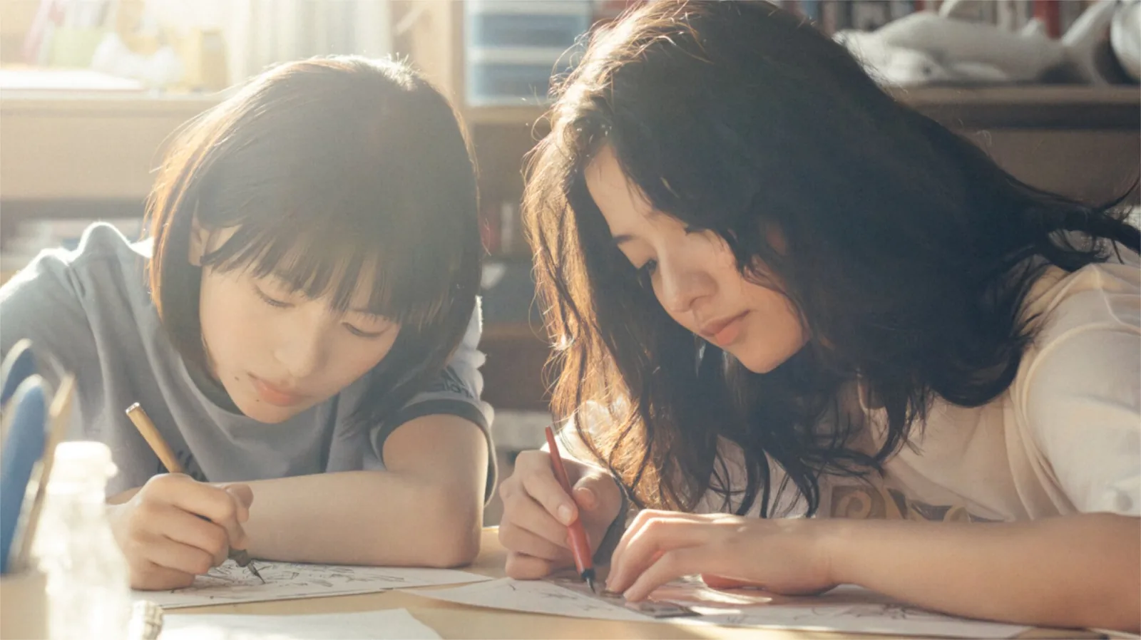 窓から差し込む光の下、二人が並んで寝そべり、紙に鉛筆で絵を描いている。