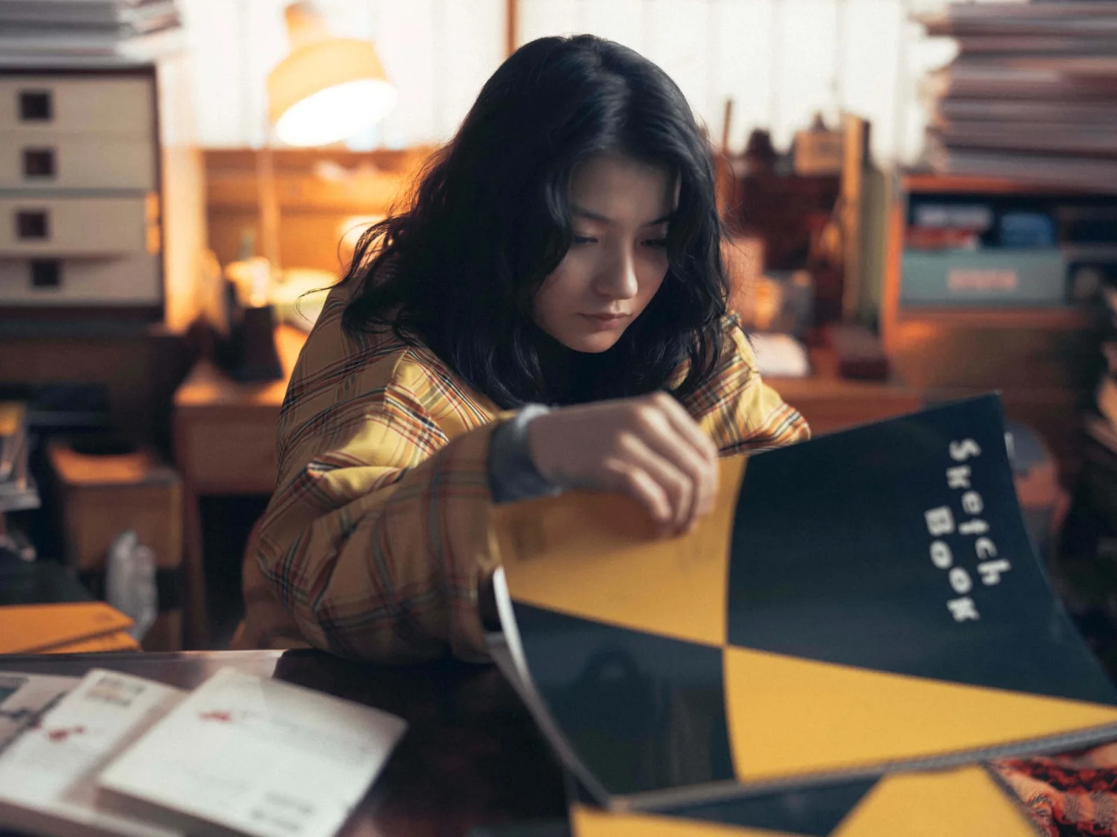 薄暗い部屋で、黄色いチェックの服の人物が、黄色と暗い色に分かれた「Sketch Book」の表紙を開いている。