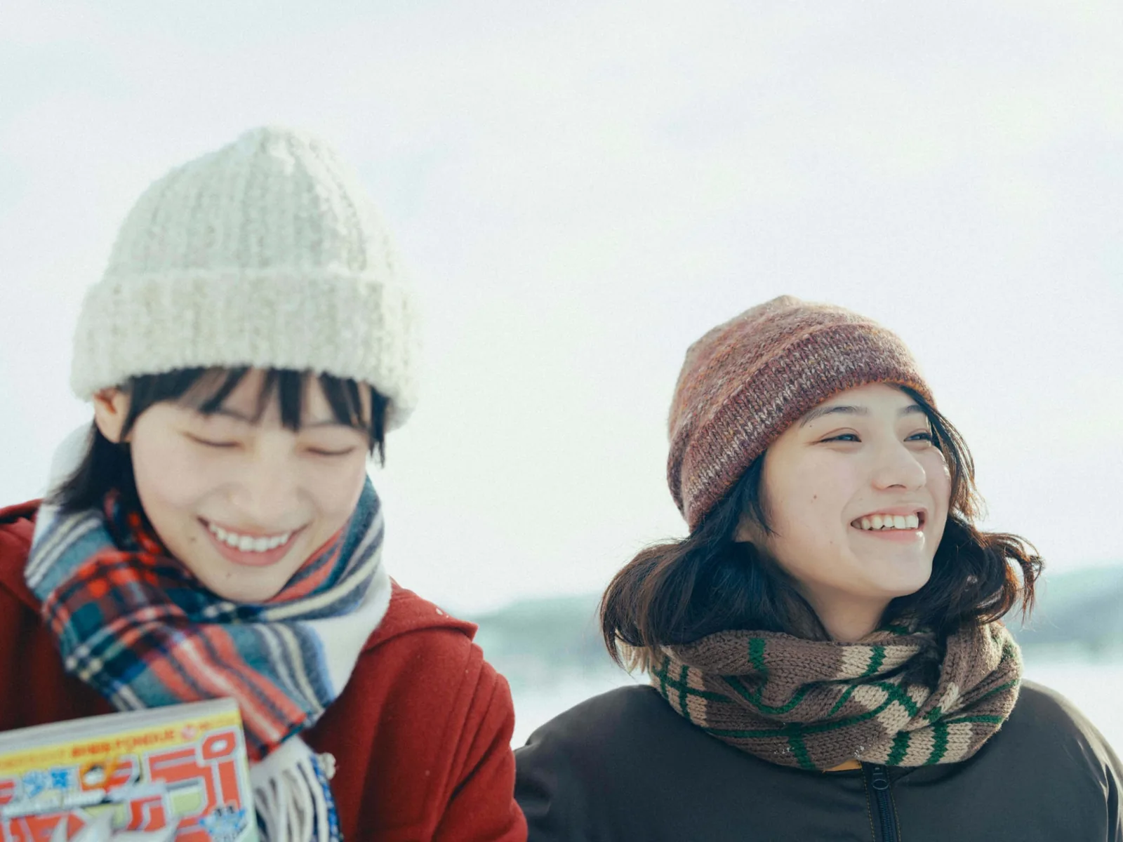 雪の屋外で、ニット帽とマフラーの二人が笑っている。左の人物は漫画雑誌を抱えている。