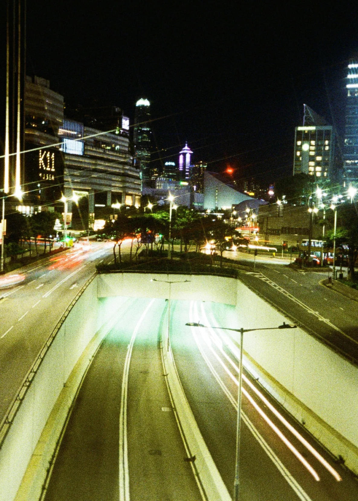 夜の市街地の道路に白と赤の車の光跡が伸び、奥に明かりのついた建物が見える。