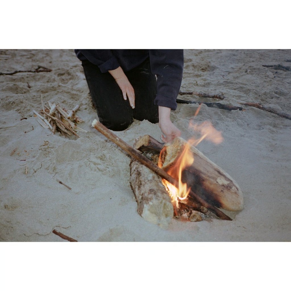 해변 모래 위 작은 모닥불 옆에서 손을 뻗고 있는 사람. 잉걸불의 따스한 톤과 모래의 회색이 균형을 이룬다.