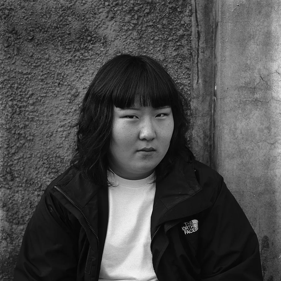 粗いセメントの壁の前に座った学生が、カメラをまっすぐ見ている。前髪をまっすぐに切りそろえ、白いTシャツの上にザ・ノース・フェイスの黒いウインドブレーカーをはおっている。