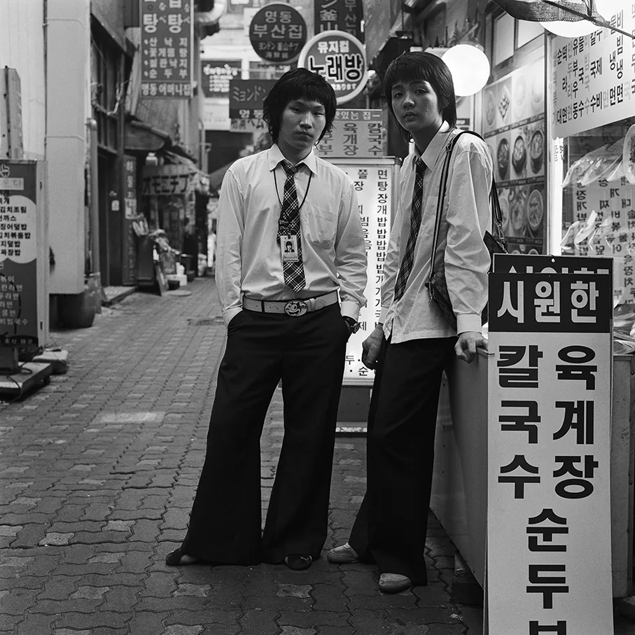 明洞の路地の食堂の前に、男子学生が二人立っている。二人とも白いシャツにネクタイを締め、左の学生は裾の広い黒のベルボトムをはいている。手前には、ユッケジャンとカルグクスを書いた縦長の立て看板がある。