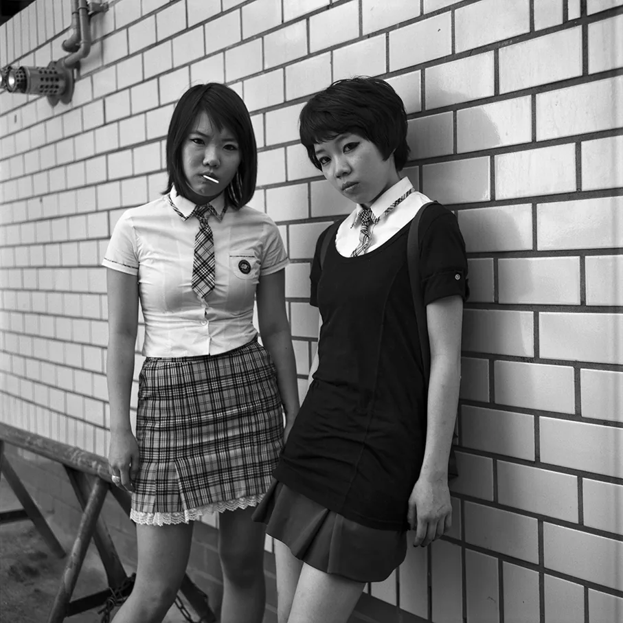 白いタイルの壁に、女子学生が二人もたれて立っている。左の学生は半袖シャツにチェックのネクタイ、裾にレースのついた短いチェックのスカート。右の学生は黒いワンピースの上にシャツの襟を出している。