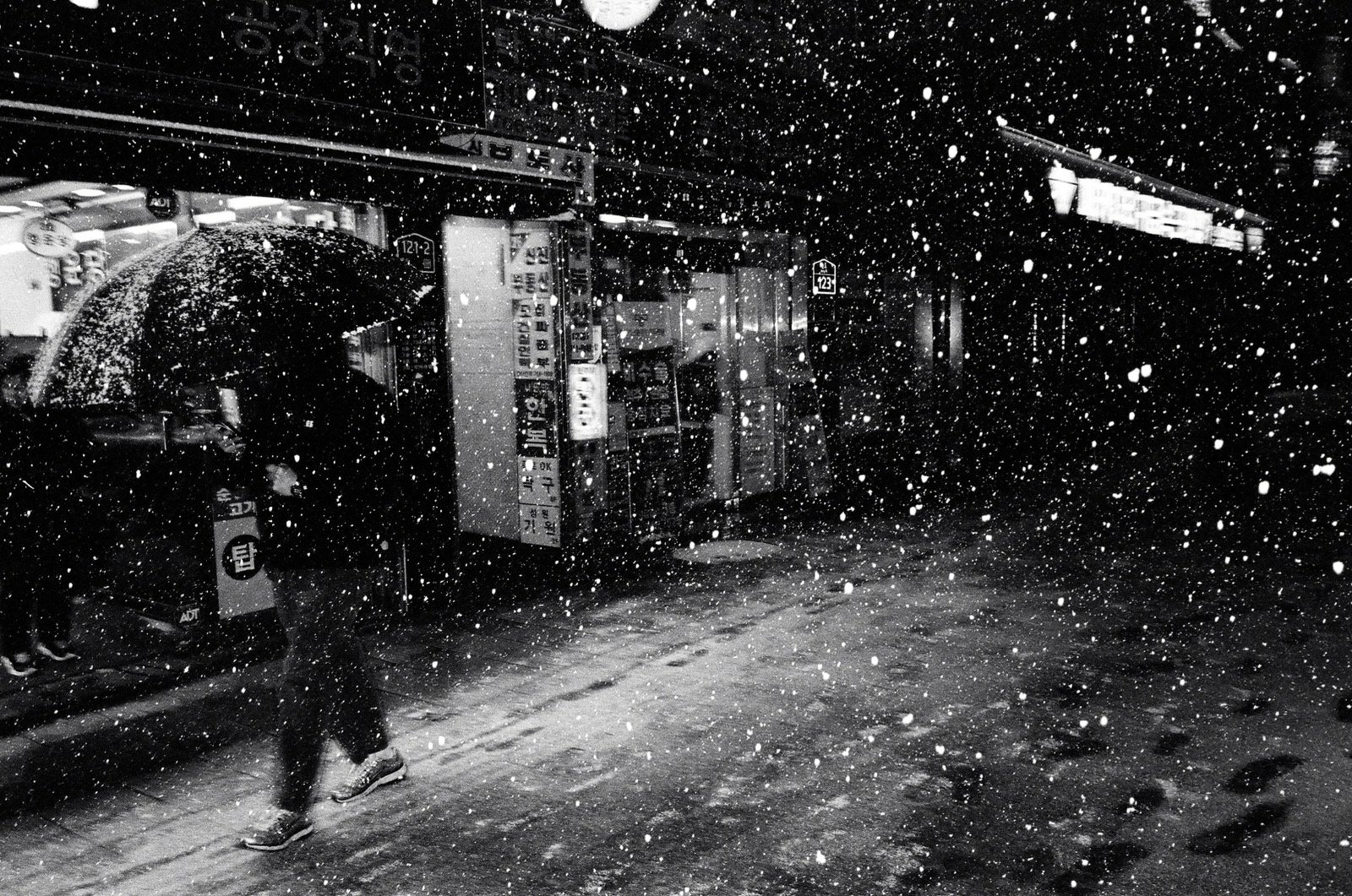 雪の降る夜、傘をさして店の前を歩く人。