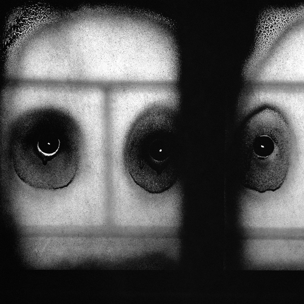 暗い画面に明るい顔のような面が浮かび、黒くにじんだ目の形が三つ見える。太い黒い帯が画面右側を縦に分け、窓枠のような淡い線が縦横に走る。