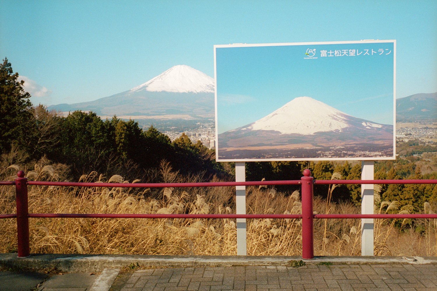 遠くに雪をかぶった富士山が見え、手前の展望台の柵の向こうに富士山の写真の看板が立っている