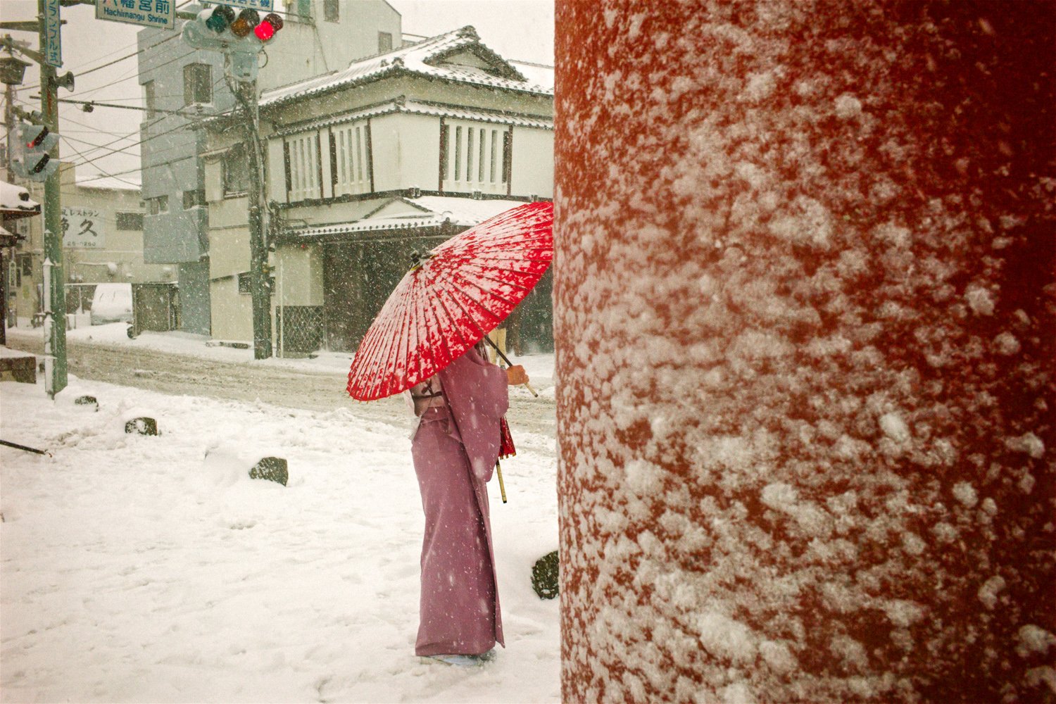 雪の積もった通りに赤い傘の人が立つ。右半分は雪のついた赤い柱がふさいでいる